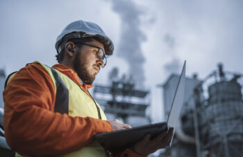 A shot of an young engineer wearing a helmet and using a laptop and hands free device during his night shirt in the oil rafinery. Engineering concept.